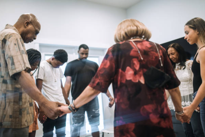 A group of people holding hands in a circle and praying together, representing faith in times of war, pandemic, and crisis.
