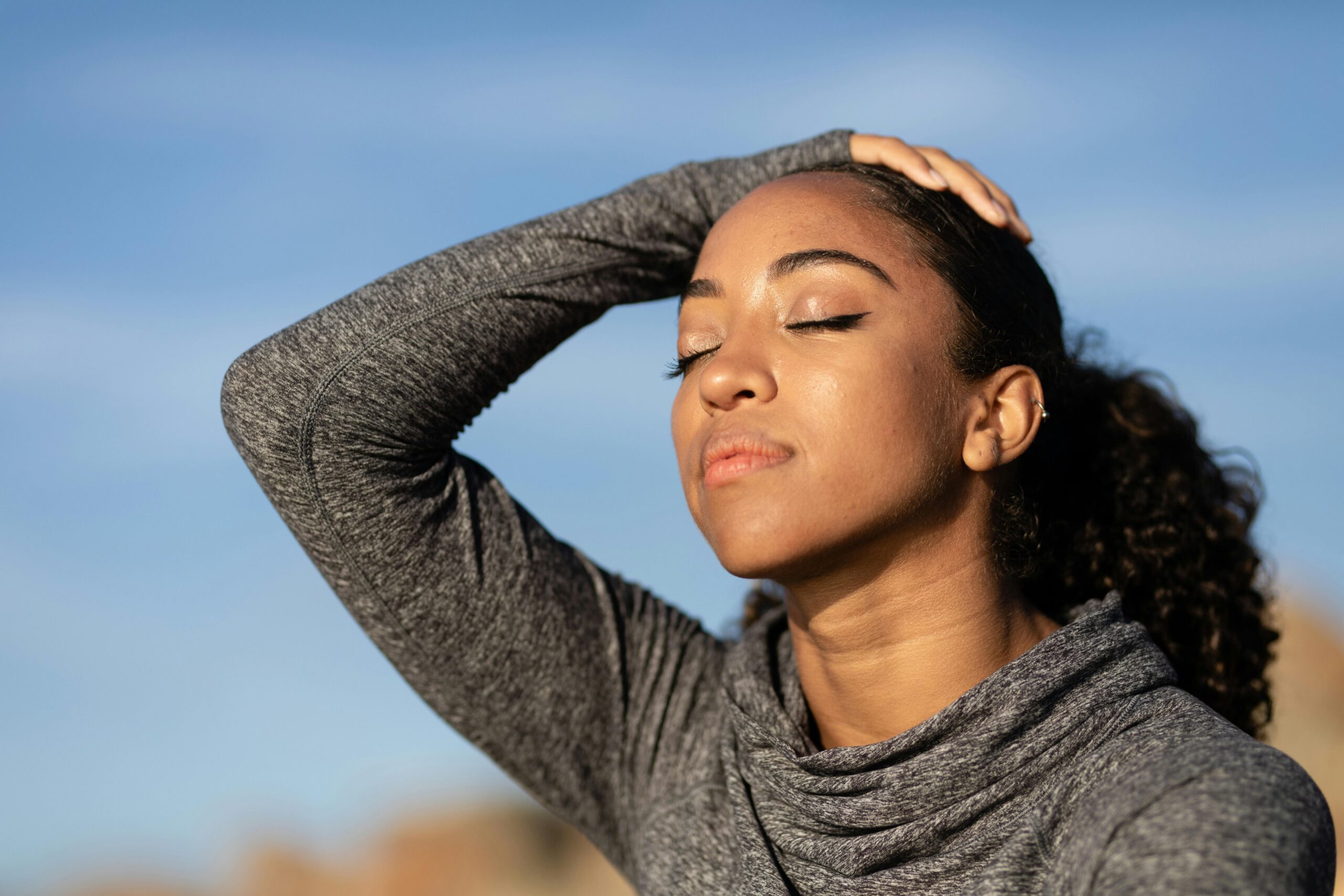A woman outdoors with closed eyes holding her head in the sunlight, practicing spiritual discernment and listening for God's voice.
